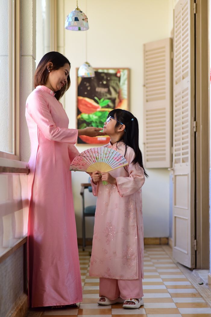 A mother and daughter in traditional pink dresses share a moment indoors, embracing Vietnamese culture.