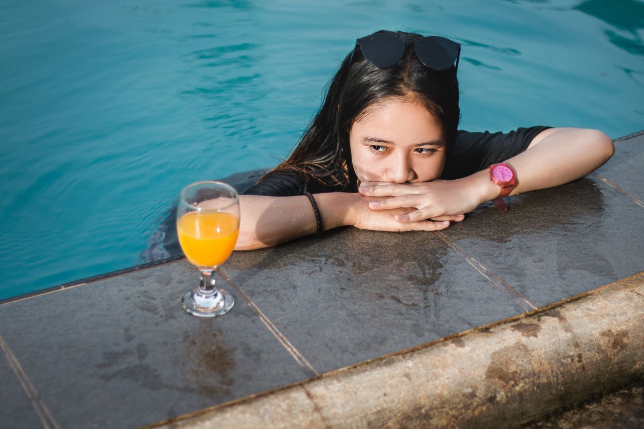 Woman relaxing in a pool with a glass of orange juice beside her, wearing sunglasses and a wristwatch.