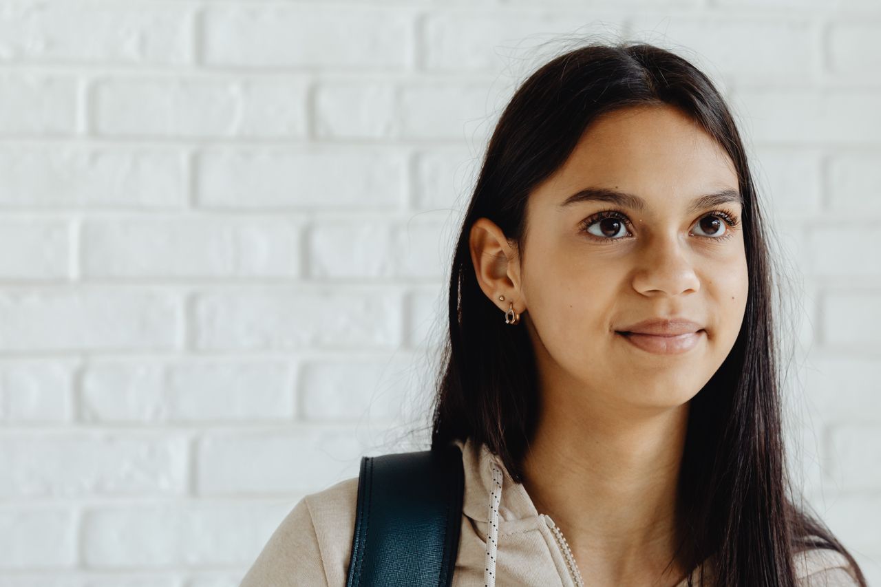 Portrait of a young woman with dark hair smiling in front of a white brick wall, wearing earrings and a backpack.