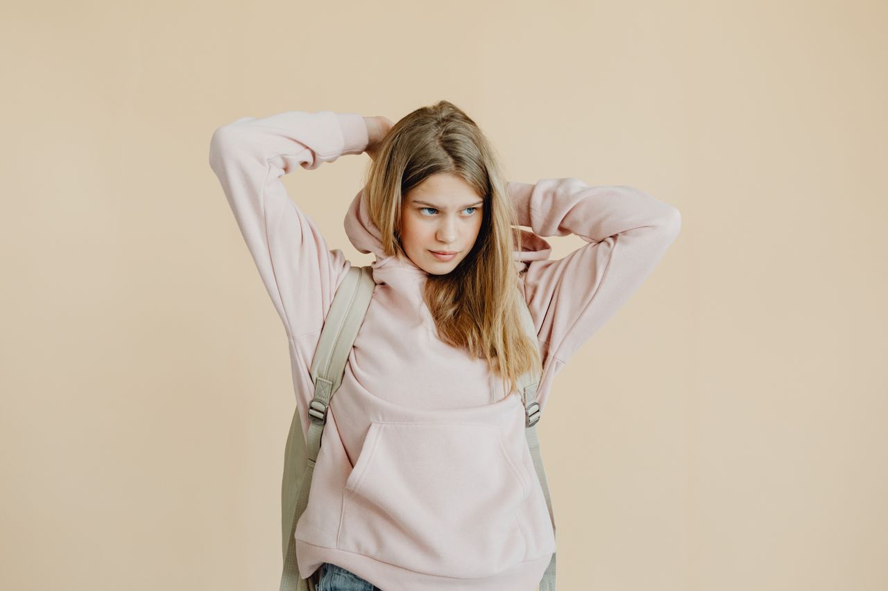 Teen girl wearing a pink hoodie and backpack, standing against a beige background.